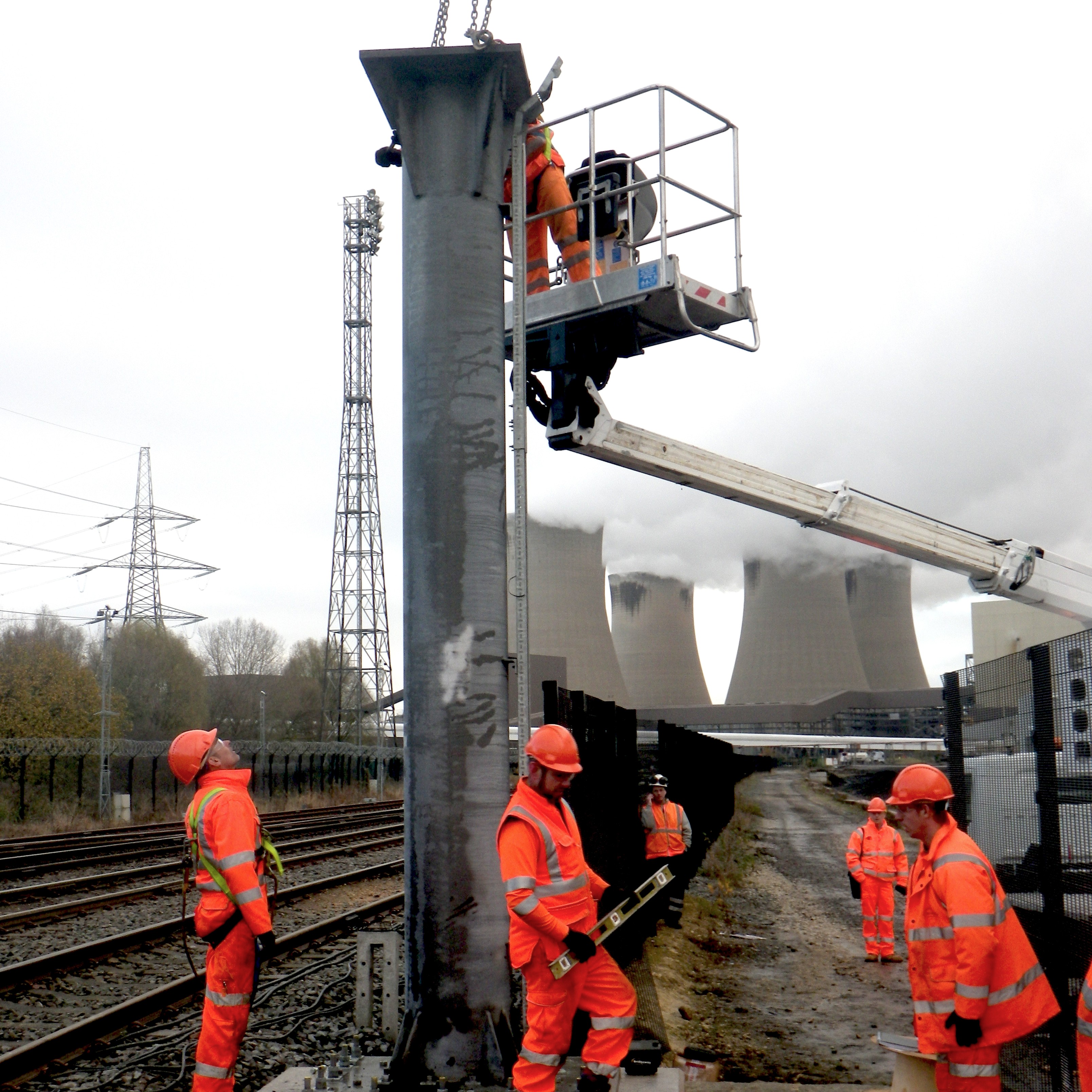 A new signal post being lifted into position beside the running lines from a mobile platform, with power station cooling towers behind.